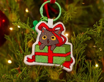 Adorno navideño bordado, ideal como llavero. Gato en la caja.