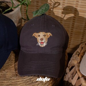 Puede incluir: Gorra de b&eacute;isbol gris oscuro con un rostro de perro sonriente bordado. El perro tiene pelaje marr&oacute;n claro y una lengua roja. La gorra tiene una visera curva y una correa de tela. La palabra "defo" est&aacute; impresa abajo a la derecha.