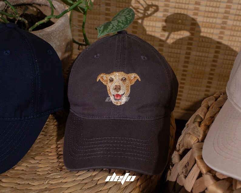 Puede incluir: Gorra de b&eacute;isbol gris oscuro con un rostro de perro sonriente bordado. El perro tiene pelaje marr&oacute;n claro y una lengua roja. La gorra tiene una visera curva y una correa de tela. La palabra "defo" est&aacute; impresa abajo a la derecha.