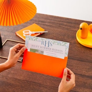 May include: An orange invitation card with the text "RHS Riley Summer Hanson" is held in an orange envelope. A matching orange lamp and notebook are on a wooden surface. A yellow tray holds a small bird-shaped object and pencils.