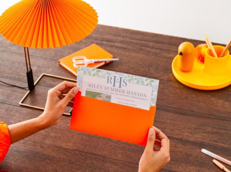May include: An orange invitation card with the text "RHS Riley Summer Hanson" is held in an orange envelope. A matching orange lamp and notebook are on a wooden surface. A yellow tray holds a small bird-shaped object and pencils.