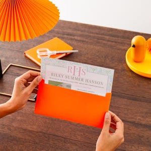 K&ouml;nnte beinhalten: Ein orangefarbener Umschlag enth&auml;lt eine Karte mit dem Text "RHS Riley Summer Hanson" auf einer Holzoberfl&auml;che. Eine passende orangefarbene Lampe und ein Notizblock befinden sich im Hintergrund, zusammen mit einem gelben Tablett mit einer Tasse und einem kleinen vogelf&ouml;rmigen Objekt.