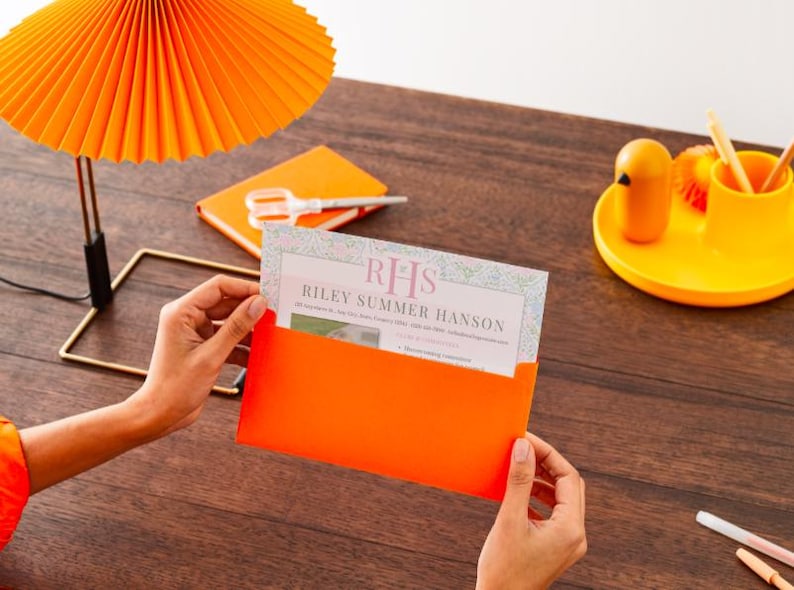 K&ouml;nnte beinhalten: Ein orangefarbener Umschlag enth&auml;lt eine Karte mit dem Text "RHS Riley Summer Hanson" auf einer Holzoberfl&auml;che. Eine passende orangefarbene Lampe und ein Notizblock befinden sich im Hintergrund, zusammen mit einem gelben Tablett mit einer Tasse und einem kleinen vogelf&ouml;rmigen Objekt.