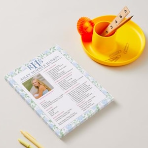 May include: A resume with the name "Riley Summer Hanson" and a photo is displayed on a white surface. A yellow desk organizer holds a pencil, ruler, and paper clips. A small orange decoration is also present.