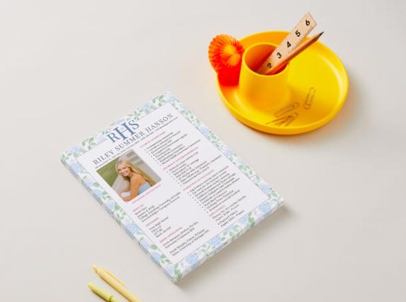 May include: A resume with the name "Riley Summer Hanson" and a photo is displayed on a white surface. A yellow desk organizer holds a pencil, ruler, and paper clips. A small orange decoration is also present.