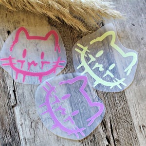 Könnte beinhalten: Drei Katzenaufkleber mit Cartoon-Design. Die Aufkleber sind in Rosa und Hellgrün gehalten und haben einen transparenten Hintergrund. Die Aufkleber befinden sich auf einer Holzoberfläche.