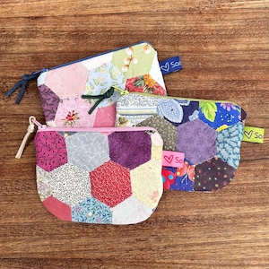 May include: Three small, colourful, patchwork zipper pouches with hexagonal patterns. The pouches feature a variety of floral and patterned fabrics in shades of pink, purple, blue, and green. Each pouch has a different coloured zip and a small tag with a heart and the word "So".