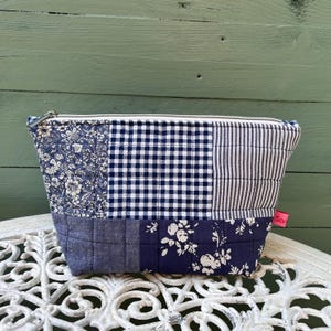 May include: A blue quilted zippered pouch with a patchwork design featuring floral, gingham, and striped patterns. The pouch is made of cotton fabric and has a zipper closure.