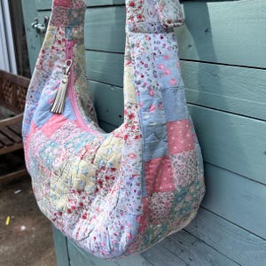 May include: A hobo-style shoulder bag constructed from a quilted patchwork fabric featuring various floral and geometric patterns in soft pastel shades of pink, blue, yellow, and white. It includes a pink zip closure and a white tassel zip pull.