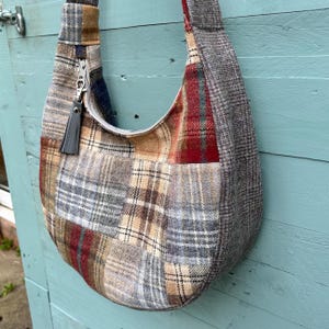May include: A patchwork shoulder bag with a curved design. The bag is made from various plaid fabrics in grey, brown, and red. It has a zip closure with a grey tassel and a matching strap.