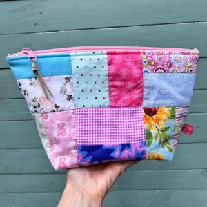 May include: A patchwork zippered pouch in various shades of pink, blue, and white. The pouch features a mix of floral, polka dot, and geometric patterns. The zipper is pink, and the pouch has a small tag that says "So".