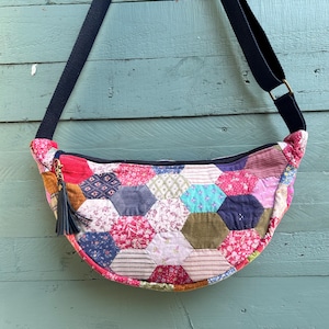 May include: A patchwork crossbody bag with a black strap. The bag is made from hexagonal fabric pieces in various floral and patterned prints. A dark blue tassel hangs from the zipper. The bag's strap is adjustable.