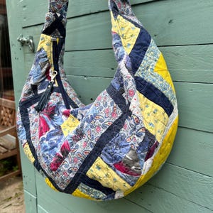 May include: A hobo-style shoulder bag constructed from quilted fabric featuring a patchwork design of yellow, navy blue, and multi-colored paisley floral patterns. The bag includes a dark blue zipper closure with a black tassel pull attached.