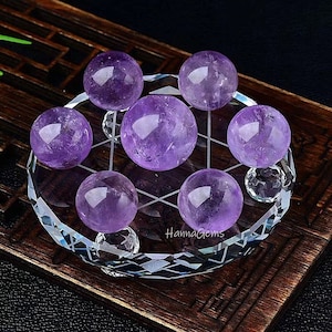 May include: Seven amethyst spheres arranged on a clear crystal base with a grid pattern. The base is on a wooden surface. HannaGems is printed on the base.