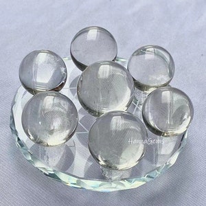 May include: Seven clear quartz crystal spheres arranged in a circle on a clear glass stand. The spheres are smooth and polished, and they reflect light beautifully.