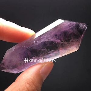 Pode incluir: Cristal de ametista translúcido e roxo com o texto "Hanna Gems" na superfície. O cristal é segurado entre os dedos contra um fundo escuro. O cristal tem uma forma polida e facetada.