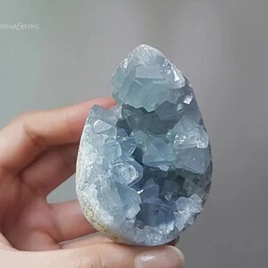 以下が含まれることがあります： 手で持たれた、水色のセレスタイトの結晶クラスター。結晶は、粗くテクスチャ加工された外観と、きらめく結晶質の内部を持つ、ジオードのような外観をしています。HannaGemsのロゴが見えます。
