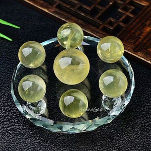 May include: Seven yellow crystal spheres of various sizes are displayed on a clear glass stand with a faceted edge. The spheres are arranged in a circular pattern on the stand.