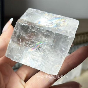 Peut inclure: Un cristal clair et rectangulaire avec des reflets irisés arc-en-ciel. Le cristal est tenu dans une main, et le texte "HannaGems" est visible à la surface. Le cristal semble être un spécimen minéral naturel.