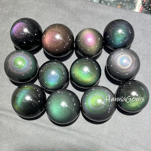 Puede incluir: Una colección de esferas de obsidiana iridiscentes. Las esferas muestran una gama de colores, incluyendo verde, púrpura y dorado, con una superficie pulida y reflectante. Las esferas están dispuestas juntas, mostrando sus patrones y texturas únicos. El texto "Hanna Gems" es visible en una esfera.