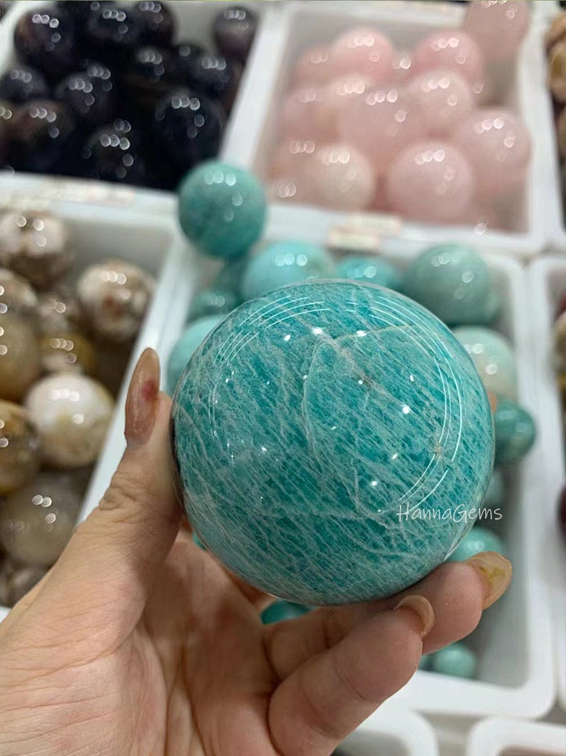 Pode incluir: Uma grande esfera de amazonita polida, de cor verde com um padr&atilde;o branco em espiral. A esfera est&aacute; sendo segurada em uma m&atilde;o.