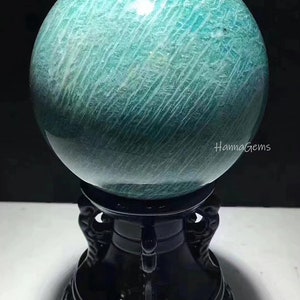 Pode incluir: Uma grande esfera de amazonita polida, de cor verde, sobre um suporte de madeira preto. A esfera tem um padr&atilde;o estriado e um acabamento brilhante. O suporte tem entalhes intrincados e uma base redonda.
