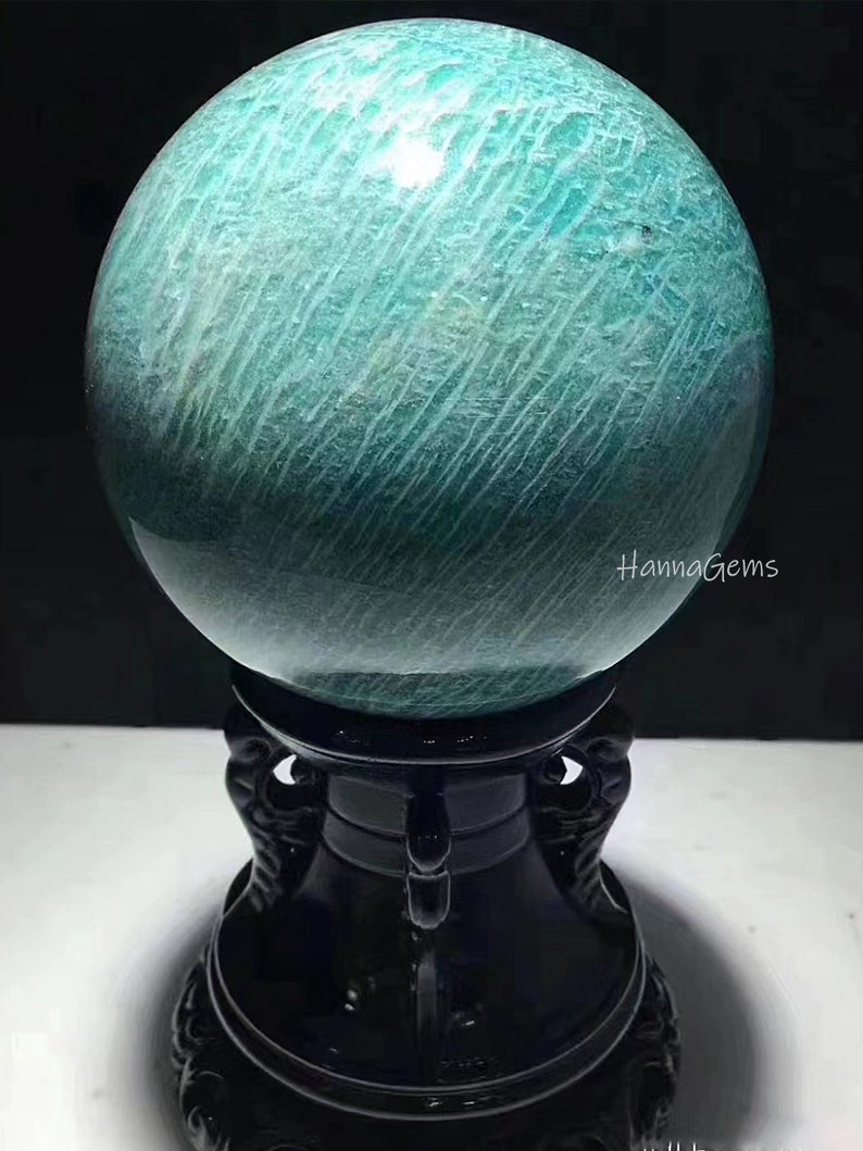 Pode incluir: Uma grande esfera de amazonita polida, de cor verde, sobre um suporte de madeira preto. A esfera tem um padr&atilde;o estriado e um acabamento brilhante. O suporte tem entalhes intrincados e uma base redonda.