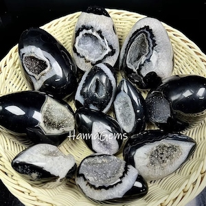 Op de afbeelding: Een verzameling gepolijste zwarte en witte agaatgeodes in een gevlochten mand. De geodes hebben verschillende vormen en maten, met zichtbare kristalformaties binnenin. De tekst "HannaGems" is zichtbaar.