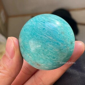 Pode incluir: Uma esfera de amazonita polida, lisa e verde. A esfera &eacute; de cor verde claro com veios brancos. A esfera est&aacute; sendo segurada em uma m&atilde;o.