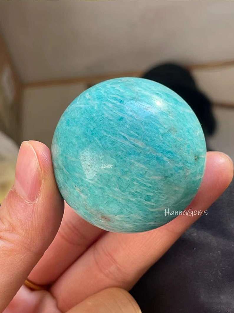 Pode incluir: Uma esfera de amazonita polida, lisa e verde. A esfera &eacute; de cor verde claro com veios brancos. A esfera est&aacute; sendo segurada em uma m&atilde;o.
