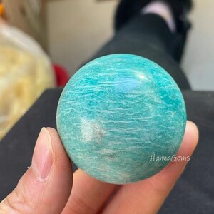 Pode incluir: Uma esfera lisa de amazonita, uma variedade de feldspato microclino, de cor verde e branca com um padr&atilde;o de redemoinho. A esfera est&aacute; sendo segurada em uma m&atilde;o.