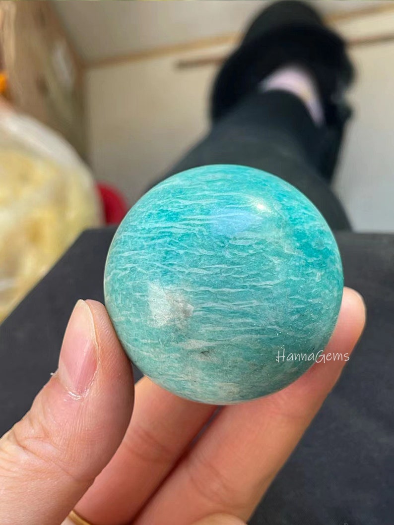 Pode incluir: Uma esfera lisa de amazonita, uma variedade de feldspato microclino, de cor verde e branca com um padr&atilde;o de redemoinho. A esfera est&aacute; sendo segurada em uma m&atilde;o.