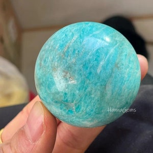 Pode incluir: Uma esfera de amazonita verde lisa e polida. A esfera tem uma cor verde clara com veios brancos. A esfera tem aproximadamente o tamanho de uma bola de golfe.