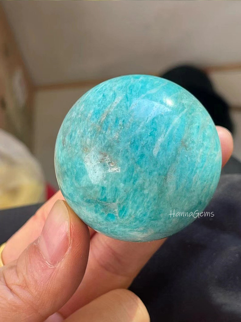 Pode incluir: Uma esfera de amazonita verde lisa e polida. A esfera tem uma cor verde clara com veios brancos. A esfera tem aproximadamente o tamanho de uma bola de golfe.