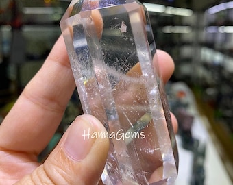 May include: A clear quartz crystal point with a faceted, pointed top. The crystal is held in a hand and has the text "HannaGems" printed on it. The crystal is transparent and has internal inclusions.