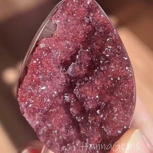 Può includere: Una gemma lucidata a forma di goccia, di colore bordeaux intenso e con un interno cristallino. La superficie brilla con minuscoli cristalli riflettenti. Il testo "Hanna Gems" è visibile. La gemma è tenuta da un supporto trasparente.