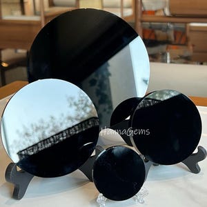 Può includere: Tre dischi neri in ossidiana lucida di diverse dimensioni su un supporto nero. I dischi sono riflettenti e mostrano un'immagine sfocata di alberi e un cielo.