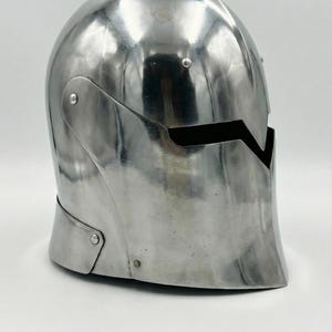 Puede incluir: Un casco pulido de color plateado, con una parte superior redondeada y una abertura para la cara. El casco tiene una superficie lisa y reflectante, probablemente de metal. Es probable que sea una pieza de armadura o un objeto decorativo.