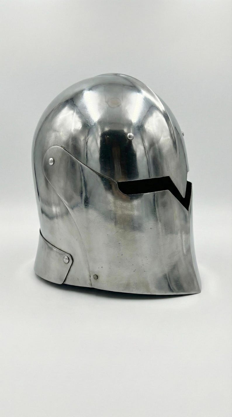 Puede incluir: Un casco pulido de color plateado, con una parte superior redondeada y una abertura para la cara. El casco tiene una superficie lisa y reflectante, probablemente de metal. Es probable que sea una pieza de armadura o un objeto decorativo.