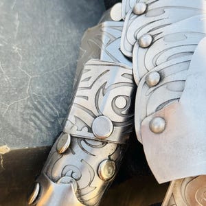 May include: Close-up of a silver metal arm guard with intricate etched designs and rivets. The guard is made of multiple overlapping plates.