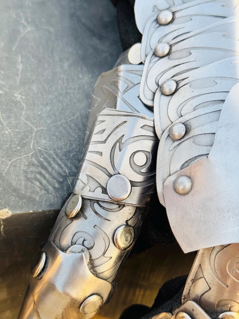 May include: Close-up of a silver metal arm guard with intricate etched designs and rivets. The guard is made of multiple overlapping plates.