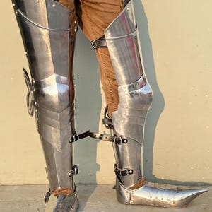 May include: A pair of silver metal leg armor with pointed toes. The armor is made of metal plates and has straps for fastening.