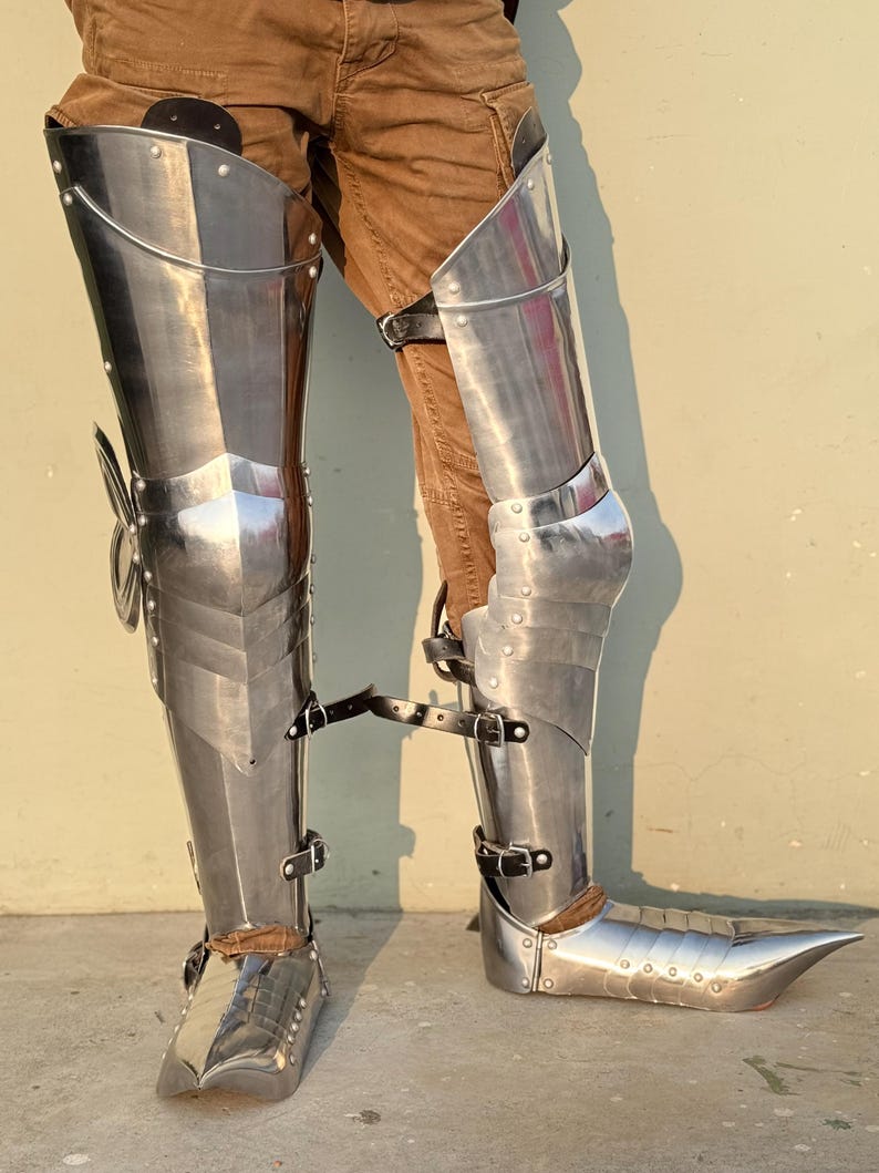 May include: A pair of silver metal leg armor with pointed toes. The armor is made of metal plates and has straps for fastening.