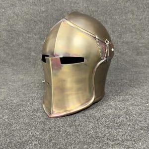 May include: A bronze-colored medieval helmet with a visor and a chain detail. The helmet has a rounded top and a pointed chin, with eye slits and a rectangular opening for the mouth. The metal has a slightly iridescent finish.