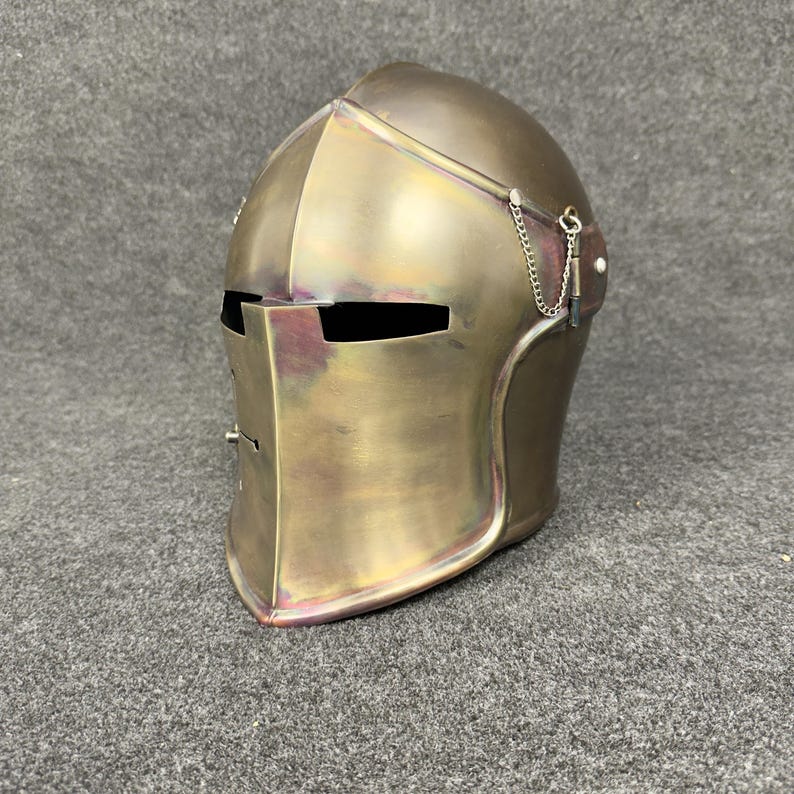 May include: A bronze-colored medieval helmet with a visor and a chain detail. The helmet has a rounded top and a pointed chin, with eye slits and a rectangular opening for the mouth. The metal has a slightly iridescent finish.