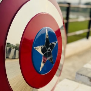 Close-up view of a Captain America shield replica. The shield is predominantly red and white, with a central blue star.  The metallic accents are polished to a high shine. The circular shield has concentric circles of red, white, and metallic silver. The blue star is detailed and reflective. This is a collectible item for fans of Marvel comics and superhero memorabilia.