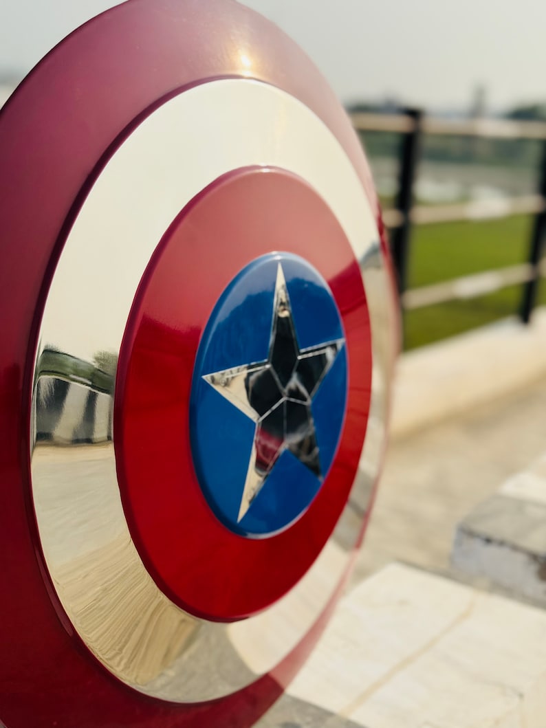 Close-up view of a Captain America shield replica. The shield is predominantly red and white, with a central blue star.  The metallic accents are polished to a high shine. The circular shield has concentric circles of red, white, and metallic silver. The blue star is detailed and reflective. This is a collectible item for fans of Marvel comics and superhero memorabilia.