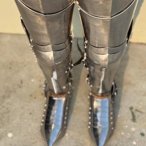 May include: A pair of silver metal shin guards with pointed toes. The shin guards are made of multiple plates that are riveted together. The shin guards are worn over brown pants.