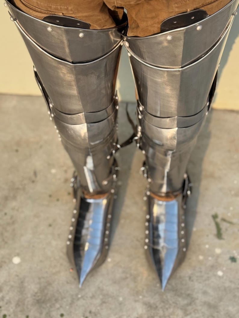 May include: A pair of silver metal shin guards with pointed toes. The shin guards are made of multiple plates that are riveted together. The shin guards are worn over brown pants.