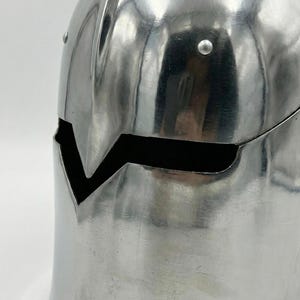 Puede incluir: Un casco plateado pulido con una distintiva abertura facial en forma de V. El casco, probablemente de metal, tiene una superficie lisa y reflectante y est&aacute; adornado con remaches. Su parte superior redondeada sugiere una est&eacute;tica medieval o fant&aacute;stica.